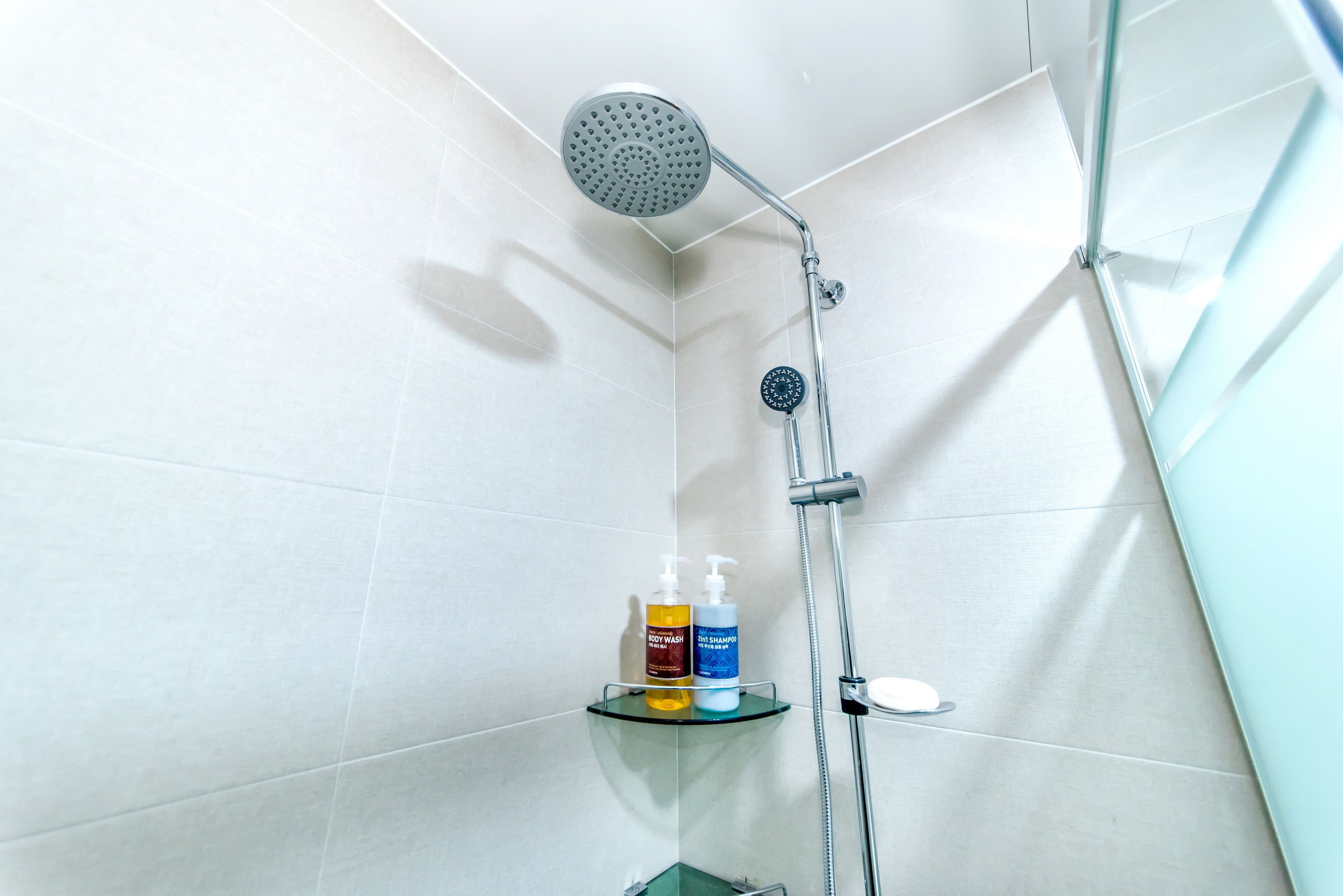A shower with a handheld showerhead and a wall-mounted faucet, a glass shelf with two bottles of shampoo, and a small mirror.