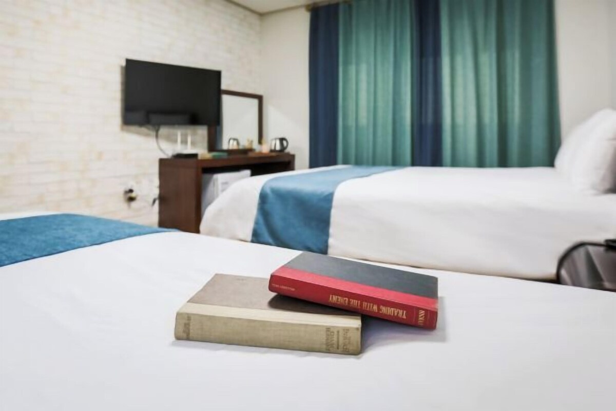 Una camera d'albergo con un letto, una TV a schermo piatto montata a parete, una piccola scrivania con lampada e una finestra con tende.
