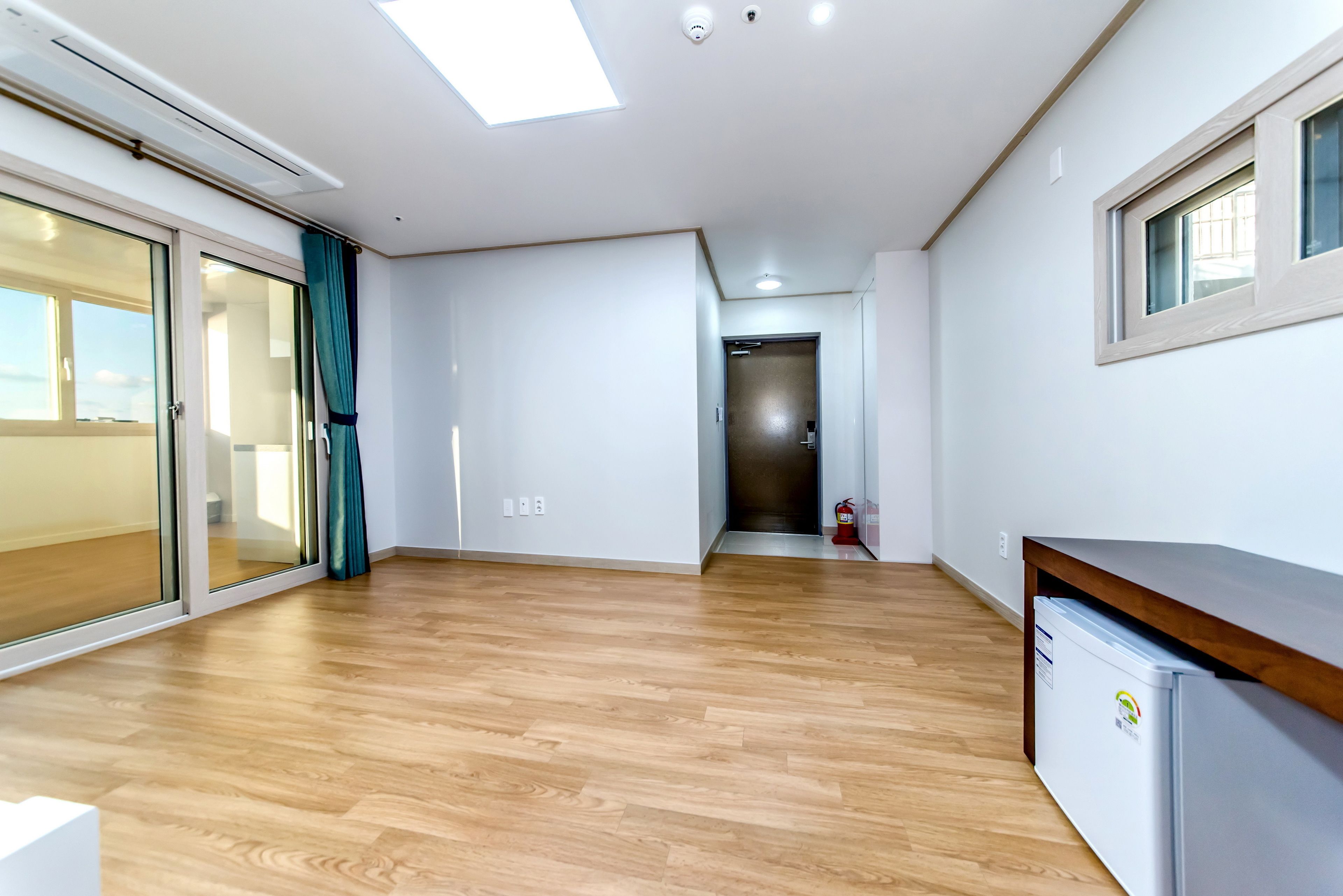 A spacious room with wooden flooring, a sliding glass door, a small refrigerator, and a ceiling light fixture.