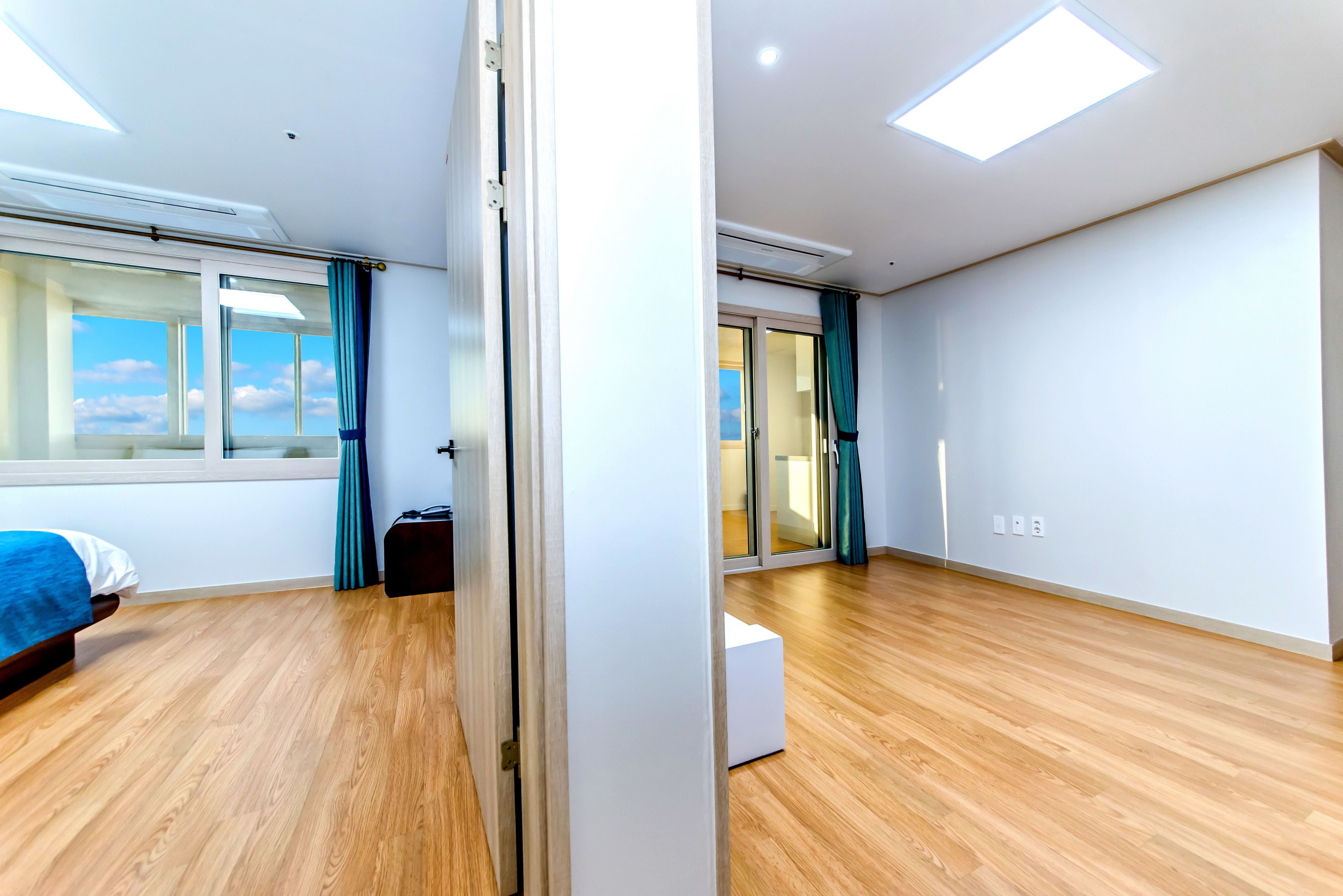 Una habitación de hotel moderna con suelo de madera, una cama, un ventanal grande con cortinas y una puerta corredera que da a otra habitación.