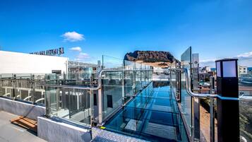 A modern glass bridge with metal railings, leading to a rocky hilltop.
