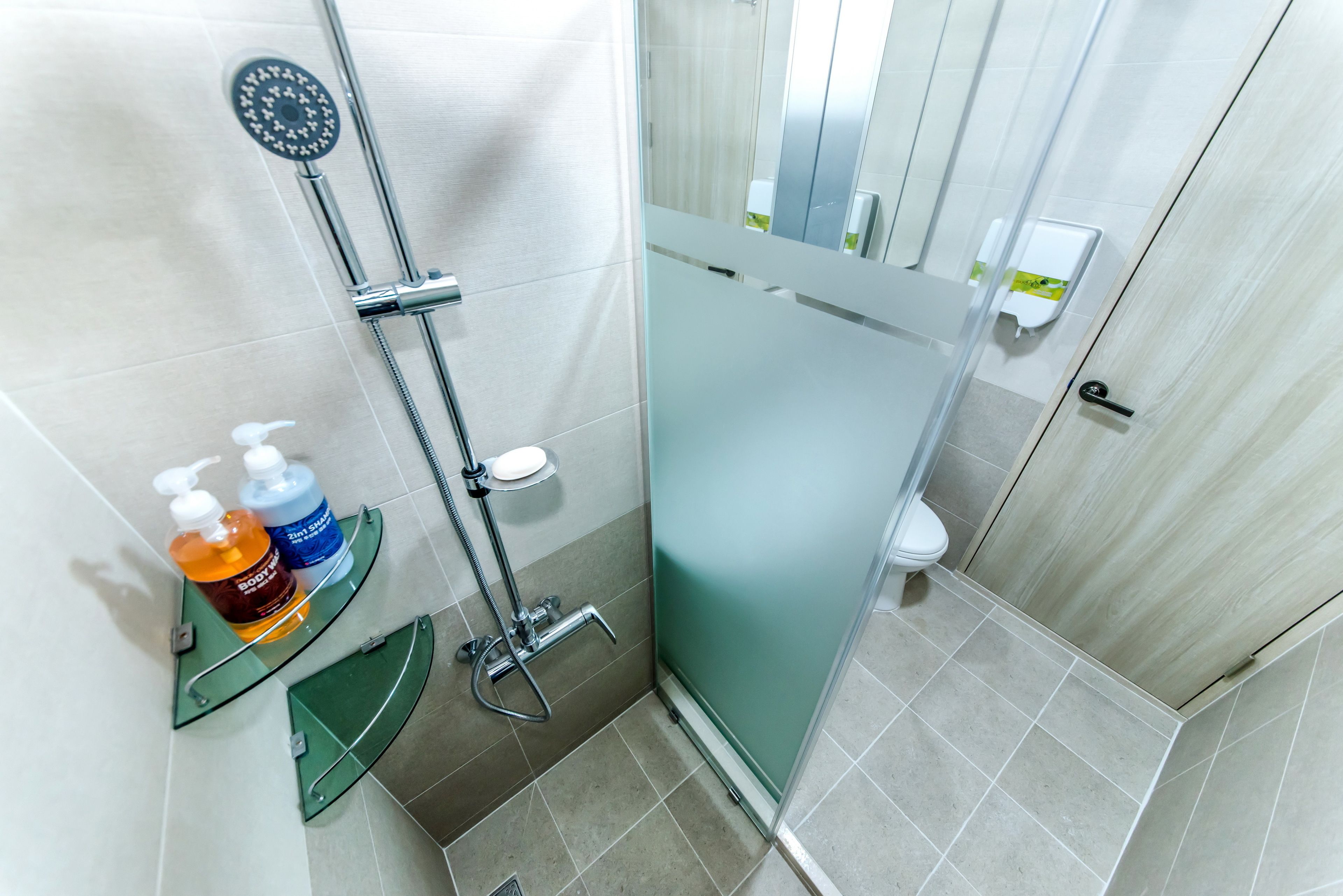 A modern bathroom with a glass shower enclosure, a handheld showerhead, and a shelf with soap dispensers.