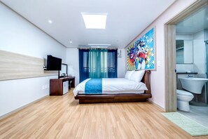 Una habitación de hotel moderna con piso de madera, una cama grande con ropa de cama blanca, un televisor de pantalla plana montado en la pared, un cuadro sobre la cama y un baño con inodoro y lavamanos.