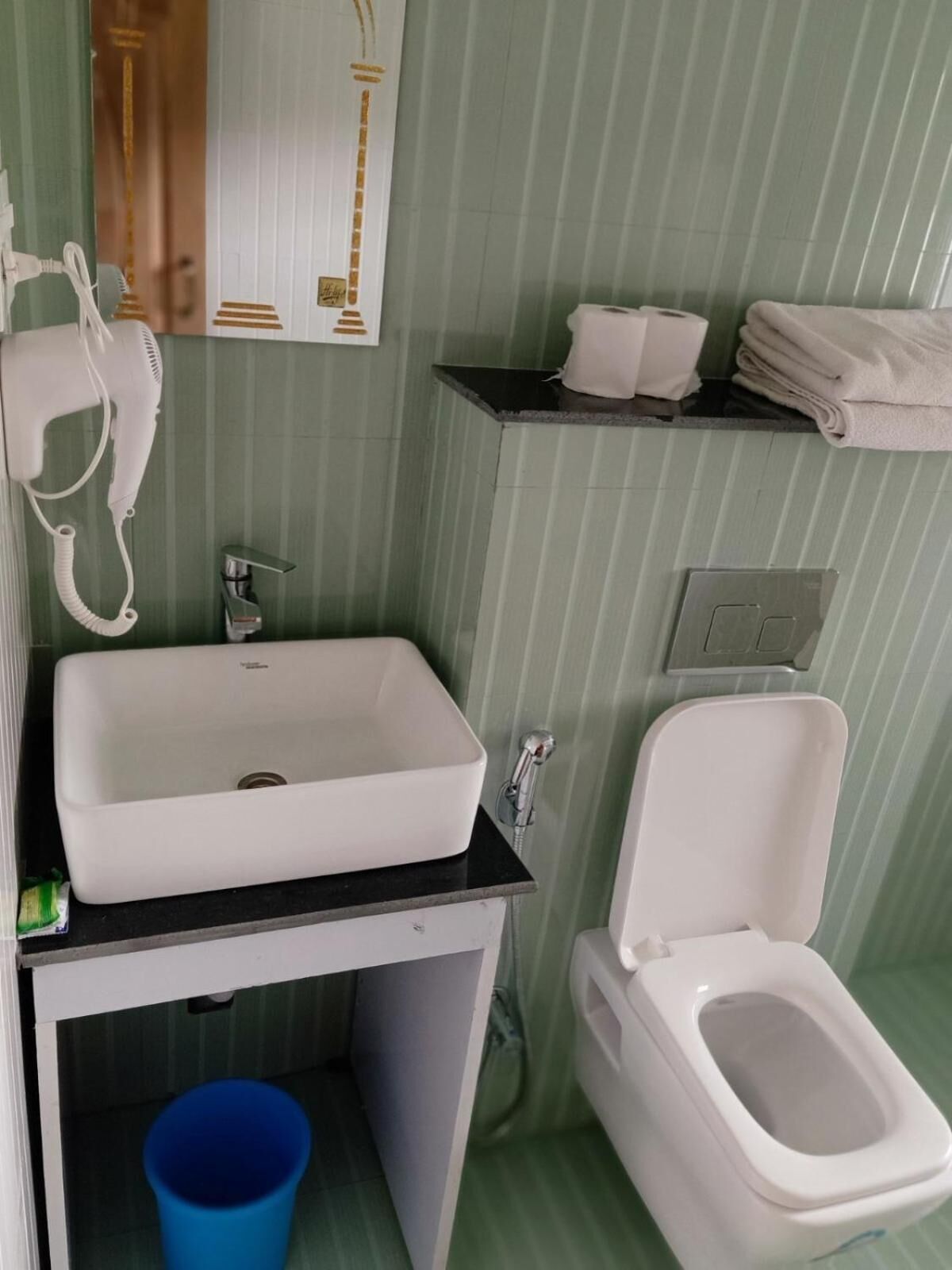 A bathroom with a white sink, a toilet, a blue bucket, and a hairdryer on the wall.