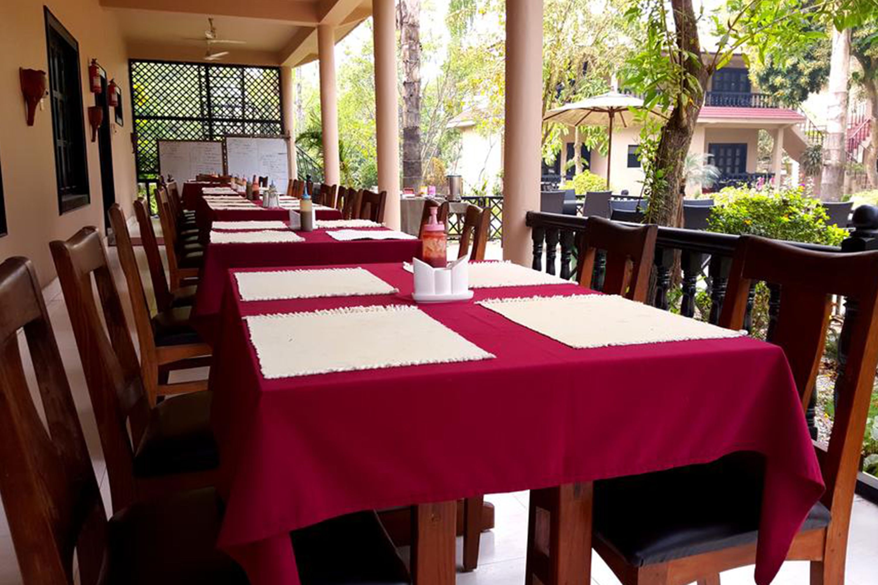 A restaurant with multiple tables covered with red tablecloths and white placemats, wooden chairs, and a view of greenery and a building in the background.