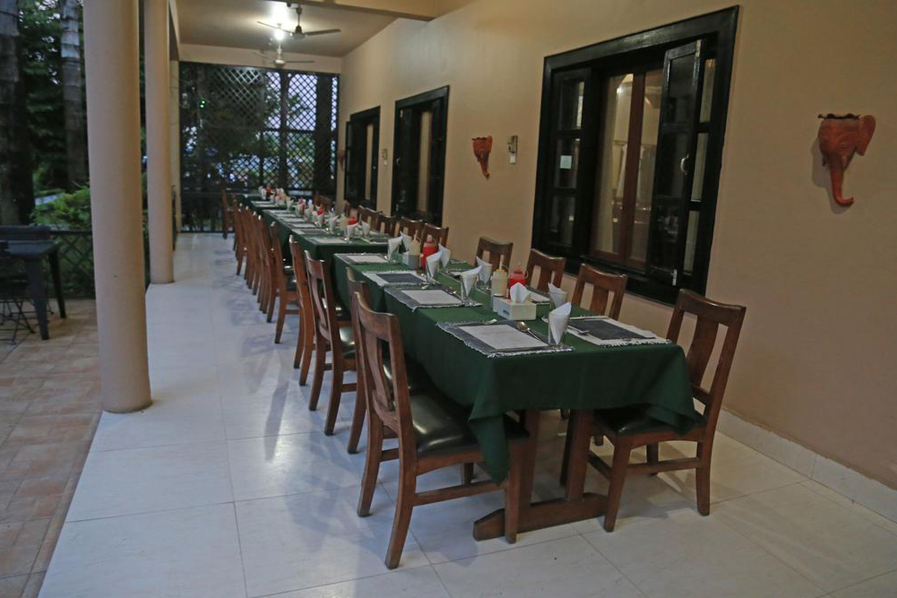A long dining table with chairs, set for a meal in a covered outdoor area with tiled flooring and a ceiling fan.