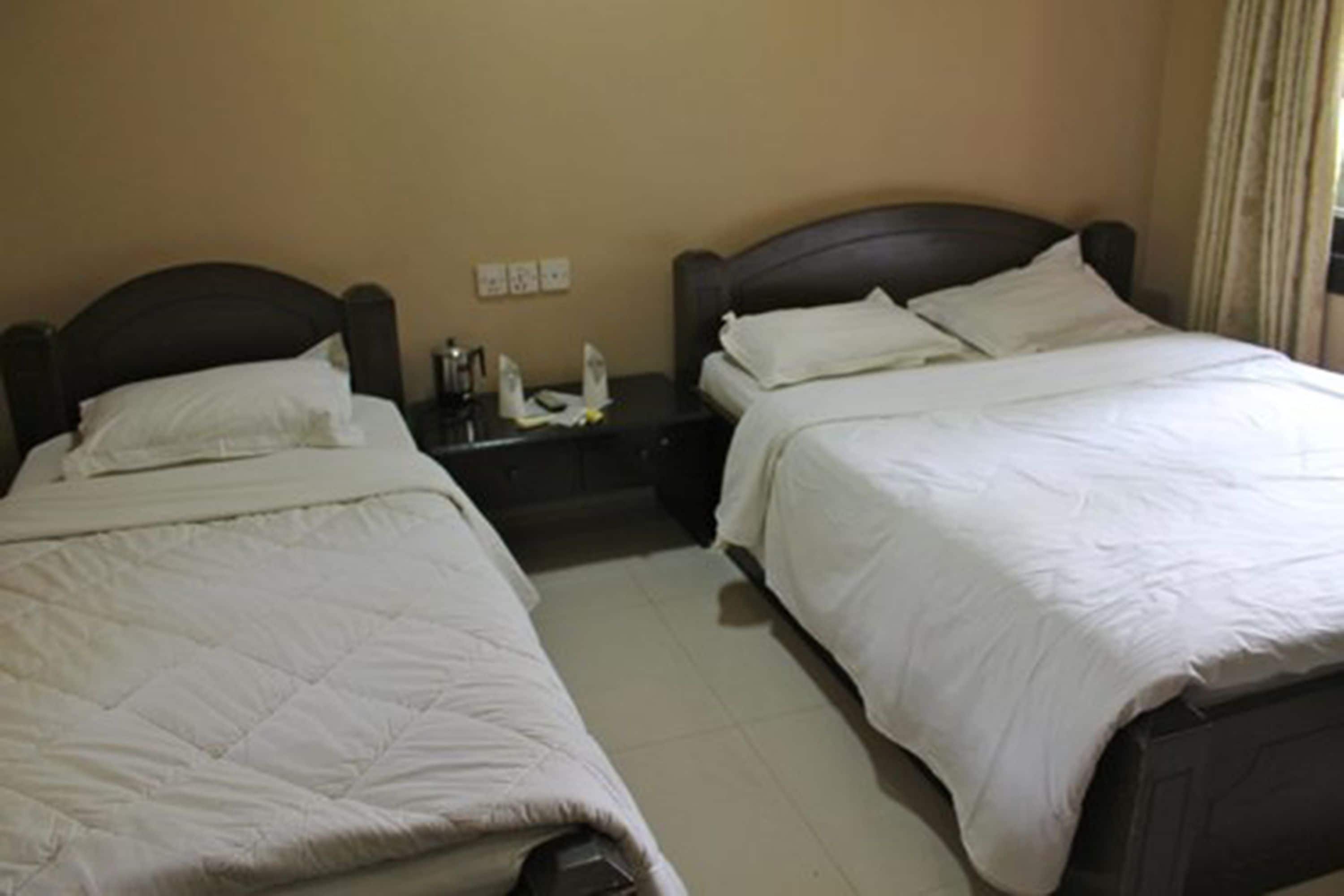 Habitación de hotel con dos camas individuales, cada una con cabecero oscuro y ropa de cama blanca. Hay una mesita de noche entre las camas con una lámpara y una botella. La habitación tiene paredes color beige y suelo de baldosas.