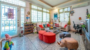 A well-lit living room with a sofa, ottomans, a coffee table, and a television. There are windows with curtains, a bookshelf, and a small table with decorative items. - Real Fun Homestay (Dongshan)