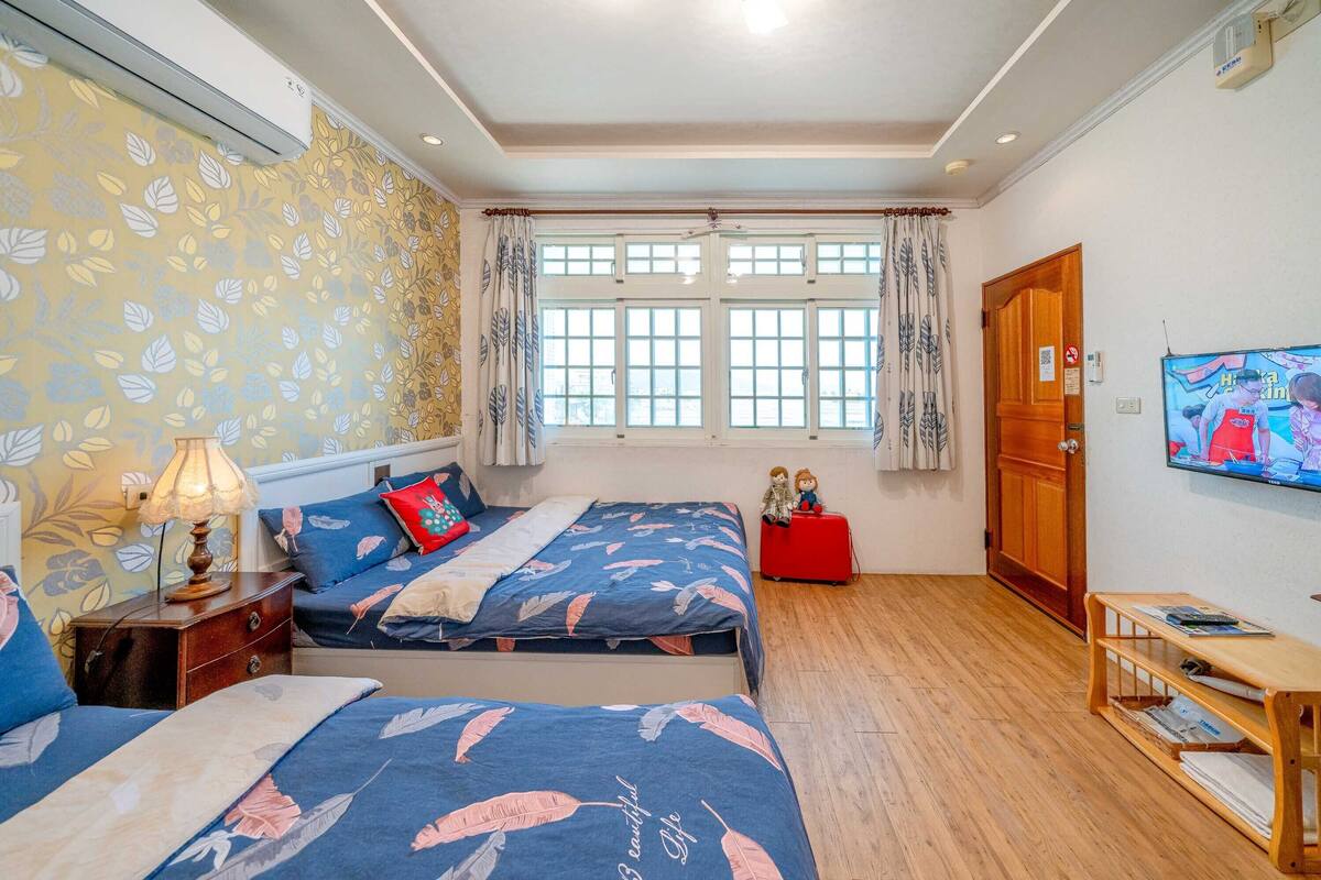 A bedroom with two beds, a TV, a wooden floor, and a window with curtains.