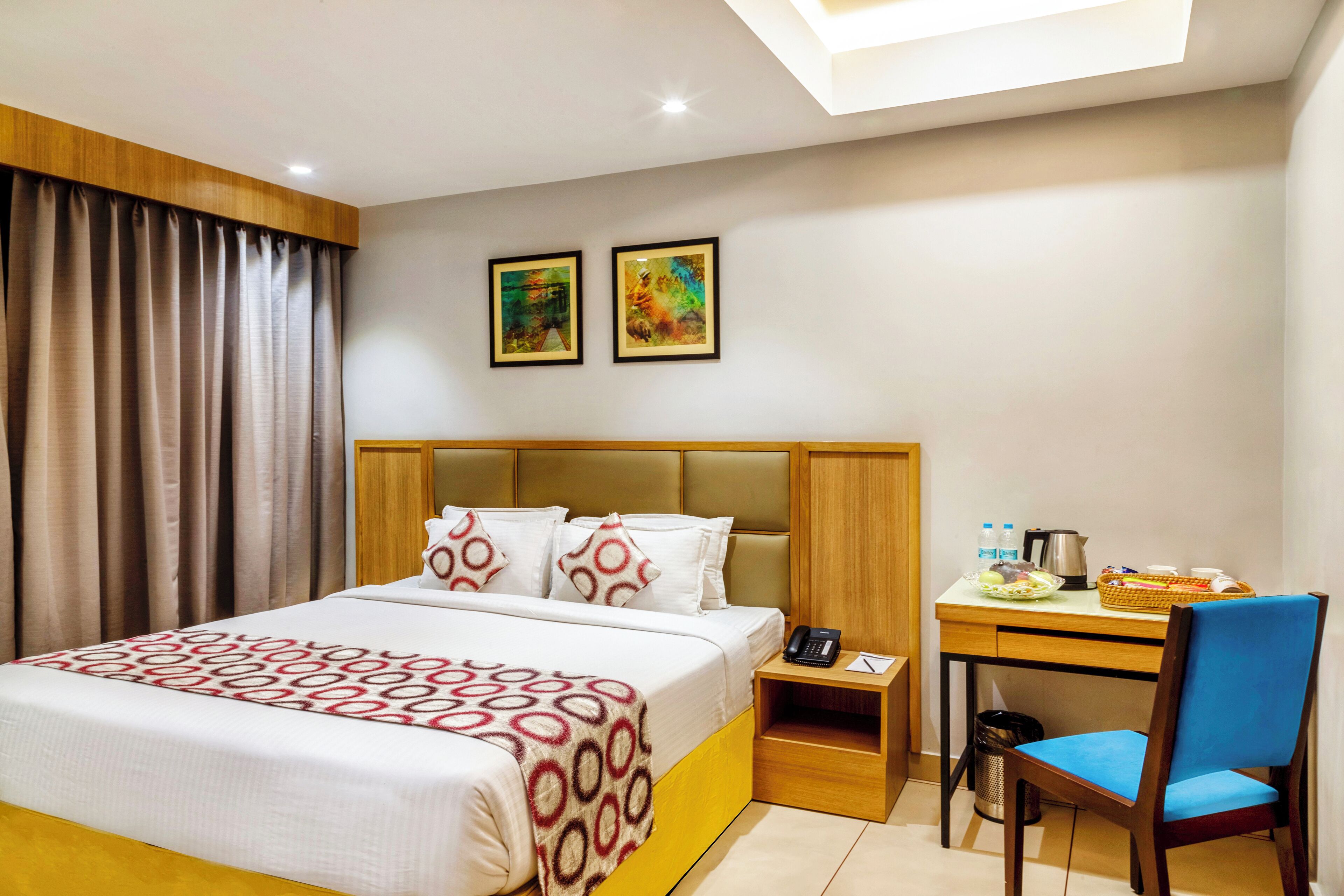 A hotel room with a bed, a desk, a chair, a nightstand, and two framed pictures on the wall.