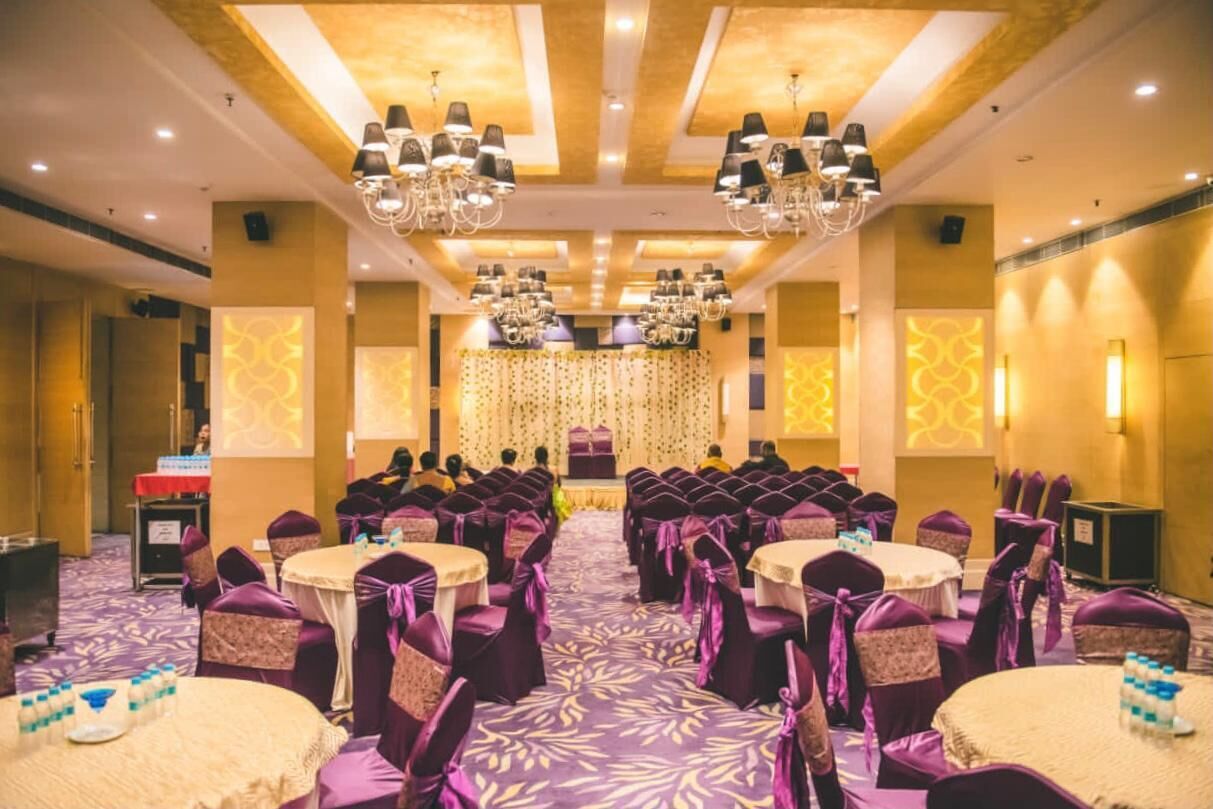 A banquet hall with round tables and purple chairs, a stage with a curtain, and chandeliers.