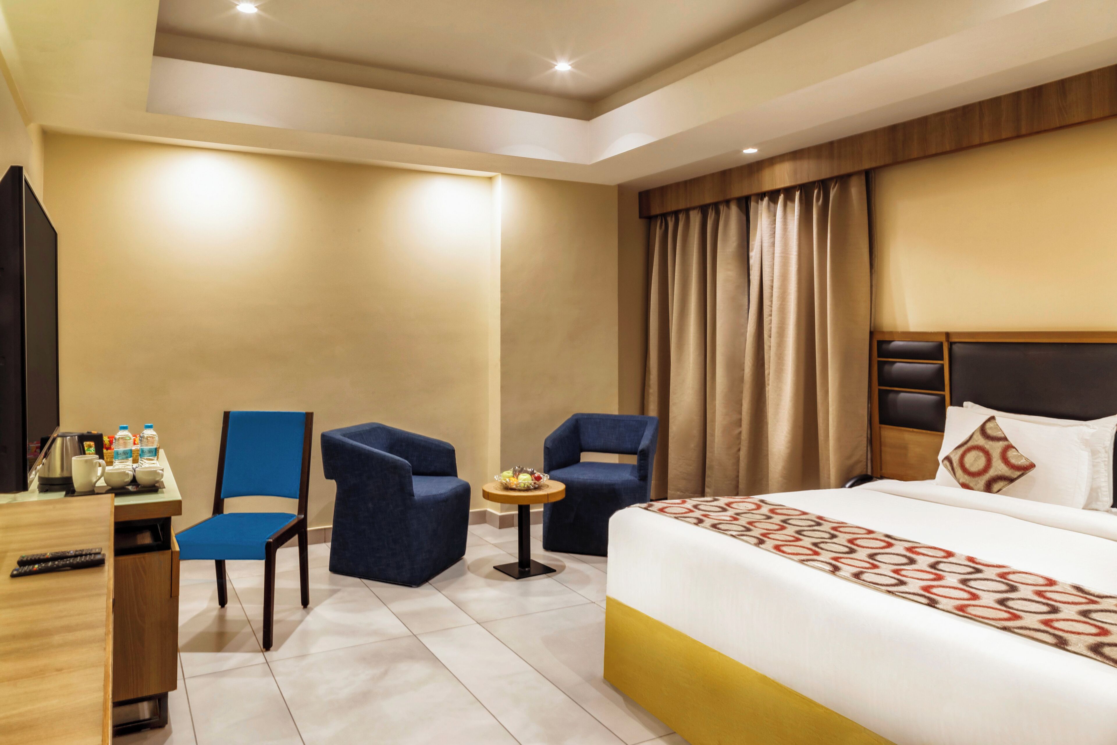 A hotel room with a bed, two blue armchairs, a small round table with a fruit bowl, a desk with a kettle and bottles, and a TV.
