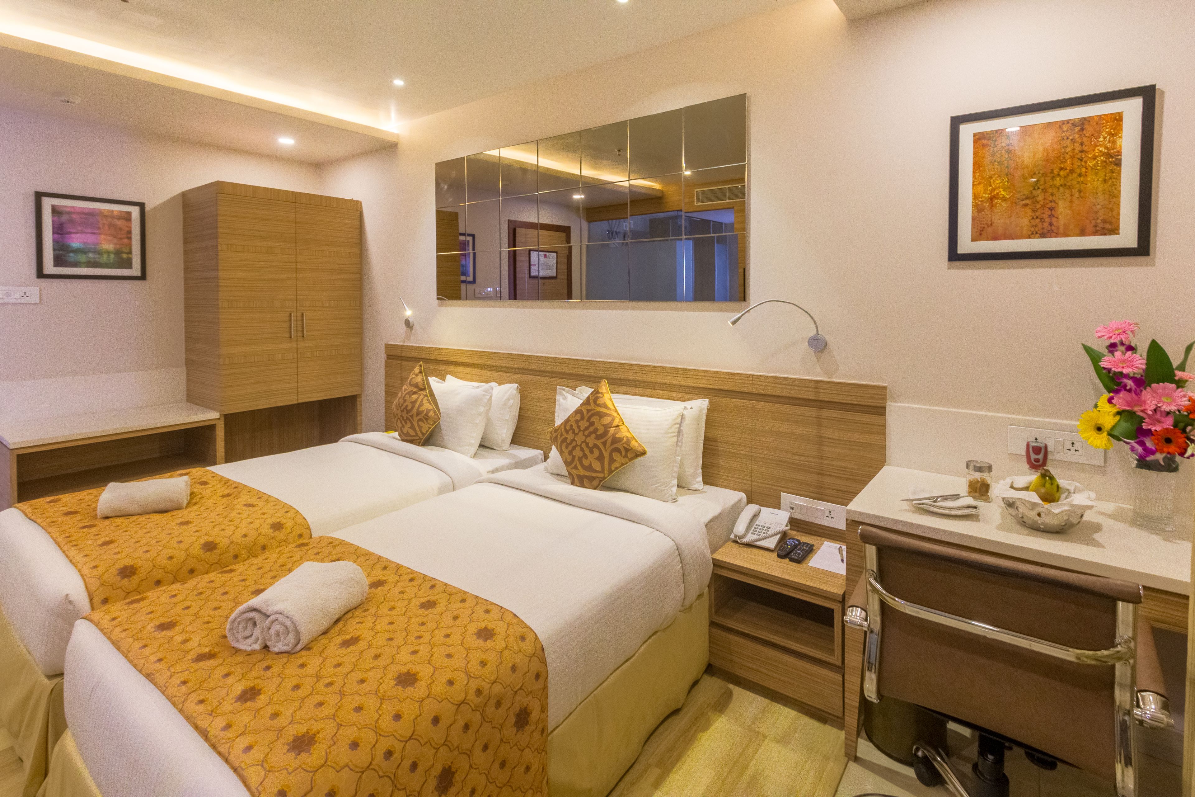 A hotel room with two beds, a wooden wardrobe, a mirror, a bedside table with a phone, and a small vase with flowers.