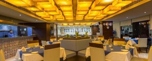 A restaurant with a modern interior, featuring a central seating area with round tables and booths, a bar area with a tiled backsplash, and a decorative ceiling with geometric patterns. - Hotel Palacio (Guwahati)