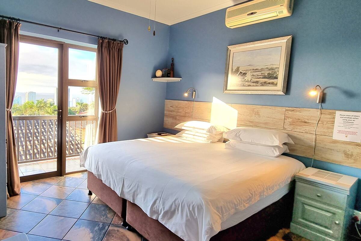 Ein Hotelzimmer mit einem Bett, einem Nachttisch, einer Lampe, einem Bild an der Wand und Blick ins Freie durch eine Schiebetür.