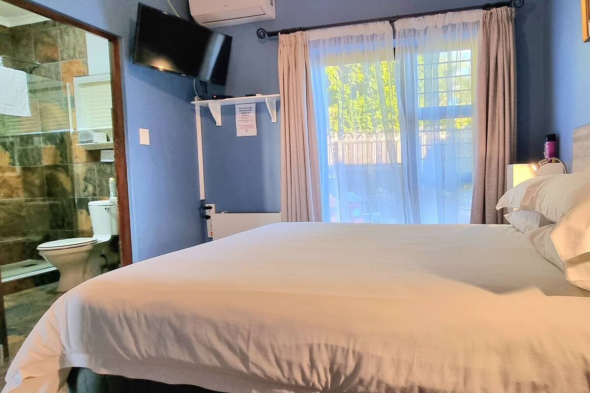 Ein Schlafzimmer mit Bett, einem an der Wand befestigten Fernseher, einem Fenster mit Vorhängen sowie einem Badezimmer mit Toilette und Dusche.