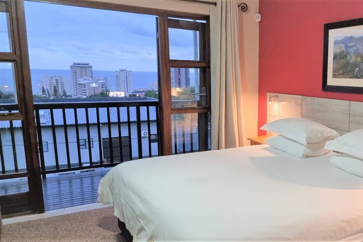 Ein Hotelzimmer mit einem großen Bett, weißer Bettwäsche, einer roten Akzentwand und Blick auf eine Stadtlandschaft vom Balkon aus.