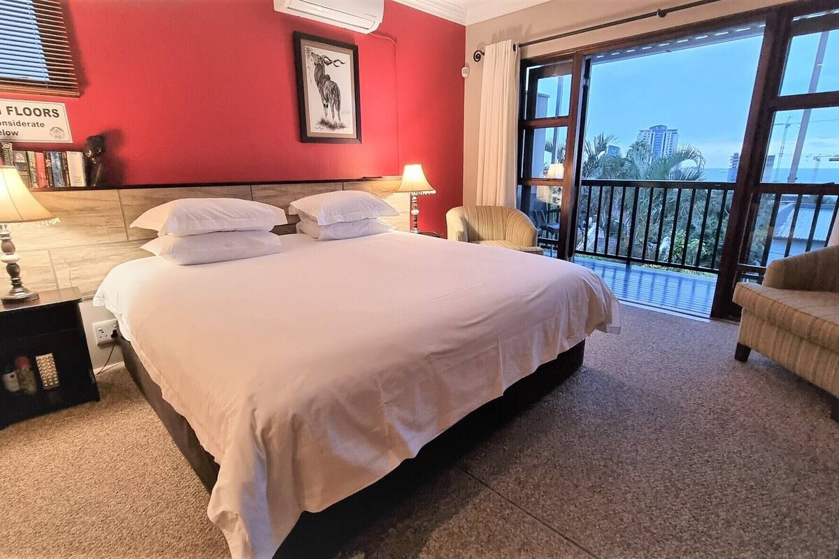Ein Hotelzimmer mit einem großen Bett, einem Sessel, einem Nachttisch und Blick auf die Stadt durch große Fenster.