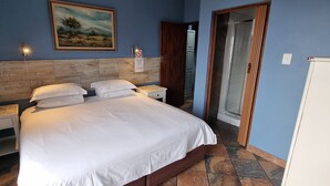 A bedroom with a bed, bedside tables, a nightstand, a painting, and a door leading to a bathroom. - Crooked Tree Cottage (Umhlanga)