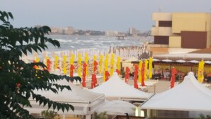 A beachside area with white tents and yellow and red umbrellas, a view of the sea, and buildings in the background. - The Premier Studio (Constanta)