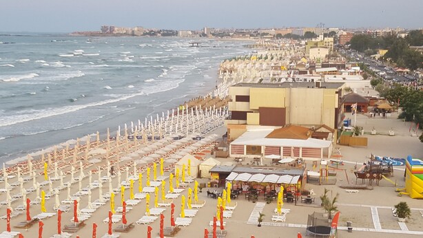 A beach with rows of sun loungers and umbrellas, a building with a flat roof, and a view of the sea and cityscape in the background. - The Premier Studio (Constanta)