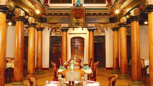 A dining area with a long table set for multiple guests, flanked by tall wooden columns, and a decorative ceiling with intricate designs. - Chidambara Vilas - A Luxury Heritage Resort (Thirumayam)