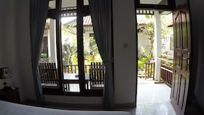A room with a bed, a window with curtains, and an open door leading to a balcony with a view of greenery and a tiled floor. - Gili Smile Bungalows (Gili Trawangan)