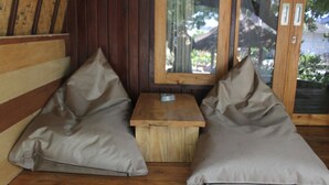 A wooden bench with two beige cushions, a small wooden table, and a glass door with a view of greenery outside. - Gili Smile Bungalows (Gili Trawangan)