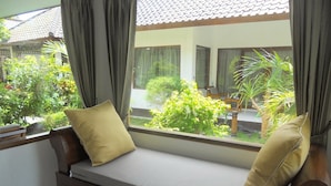 A wooden bench with cushions, a large window with curtains, and a view of a garden and building outside. - Gili Smile Bungalows (Gili Trawangan)