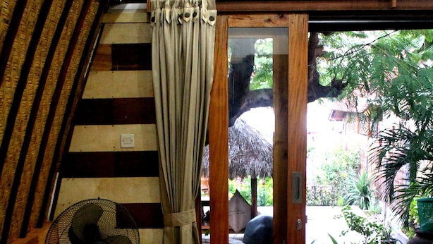 A room with a wooden floor, a bed, a fan, and a view of an outdoor area with plants and a thatched roof structure. - Gili Smile Bungalows (Gili Trawangan)