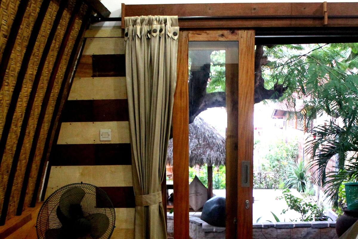 Une pièce avec un plancher en bois, un lit, un ventilateur et une vue sur un espace extérieur agrémenté de plantes et sur une structure dotée d’un toit de chaume.