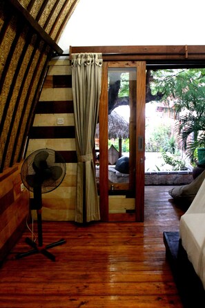 Une pièce avec un plancher en bois, un lit, un ventilateur et une vue sur un espace extérieur agrémenté de plantes et sur une structure dotée d’un toit de chaume.