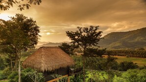 A thatched-roof hut with a wooden deck, surrounded by lush greenery and hills in the background.
