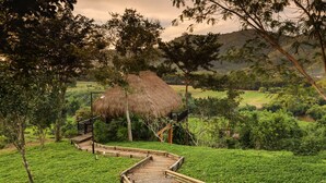 A thatched-roof structure with a wooden staircase leading up to it, surrounded by lush greenery and trees.