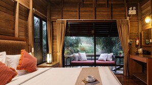 A wooden cabin room with a bed, a window with curtains, a small table with a cup and book, and a view of greenery outside.
