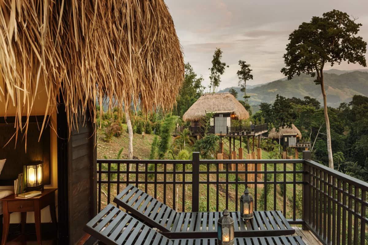A wooden deck with lounge chairs and a lantern, overlooking a thatched-roof building and lush greenery in a mountainous area.