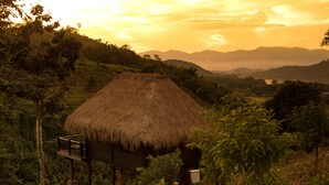 A thatched-roof hut with a wooden deck, surrounded by lush greenery and hills in the background.