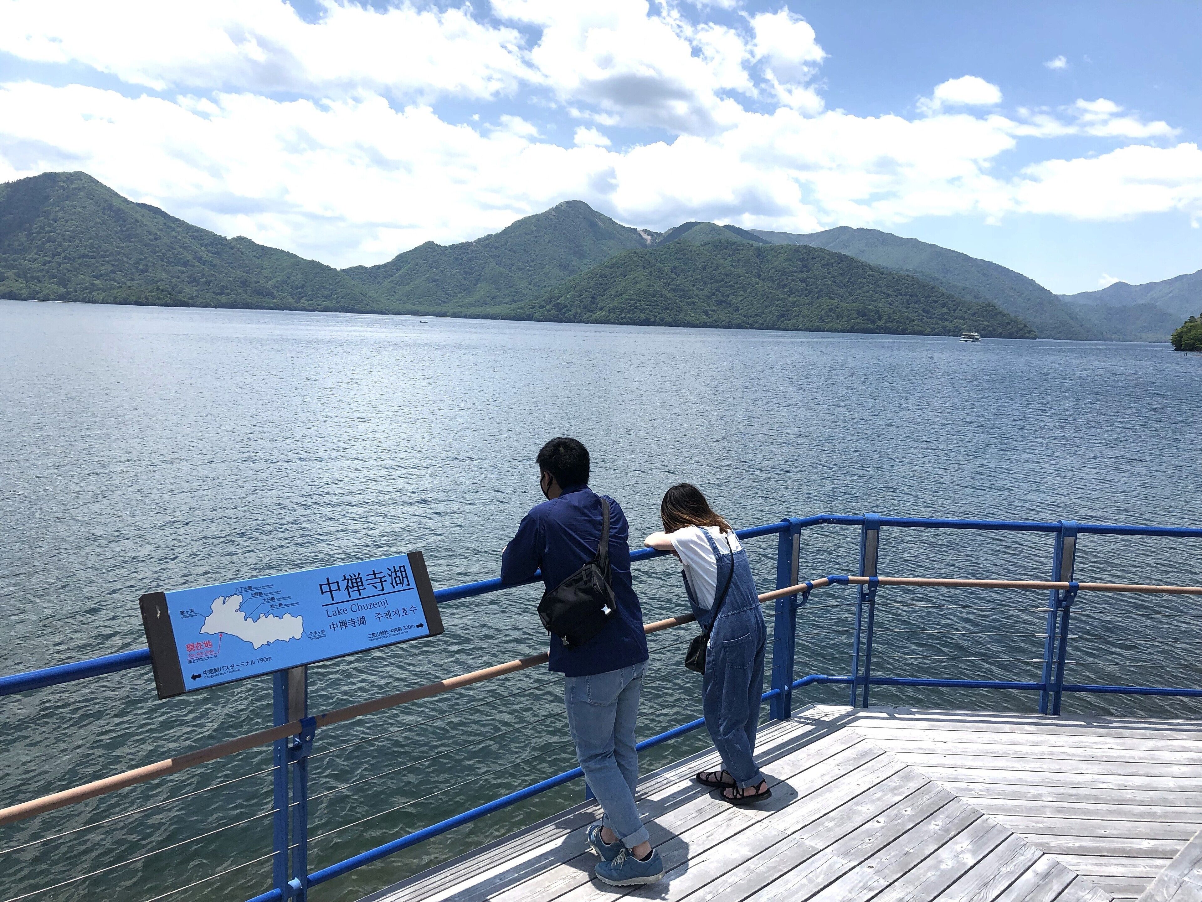 A wooden dock with a railing, a sign indicating Lake Chuzenji, and a view of the lake and mountains.