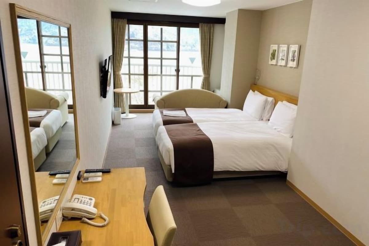 A hotel room with a bed, a desk with a telephone, a chair, a mirror, and a window with curtains.
