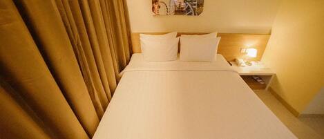 A hotel room with a bed, curtains, a nightstand, and a wall-mounted lamp.
