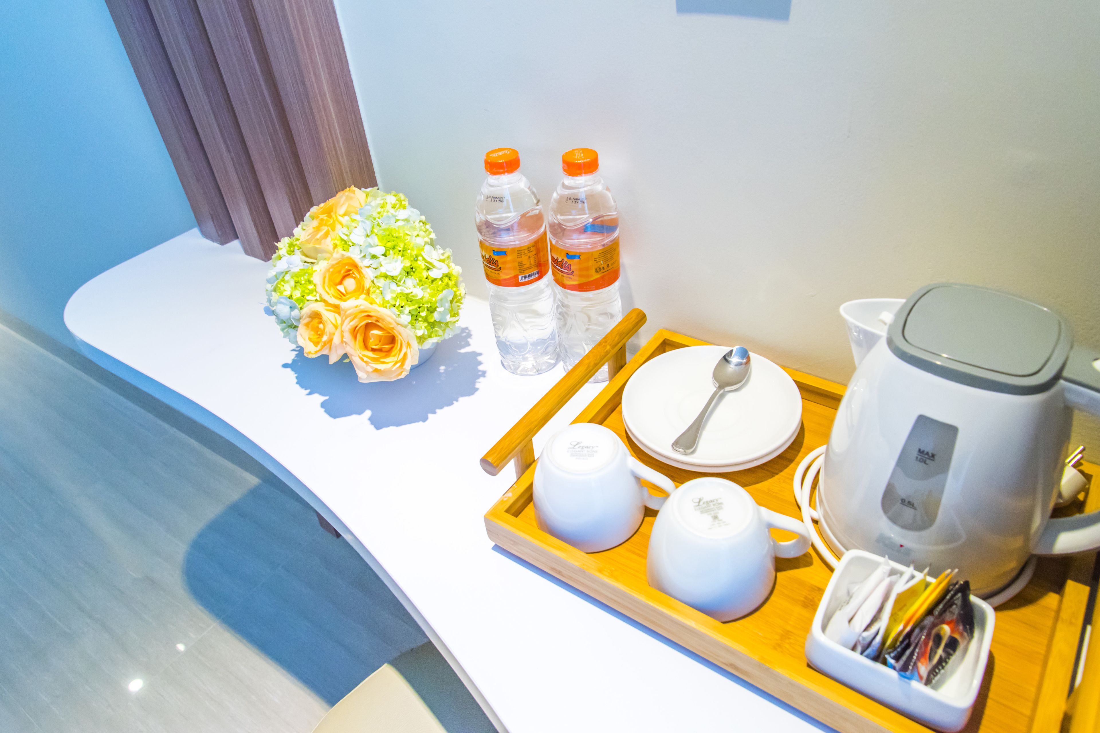 A hotel room with a wooden tray holding a tea set, two bottles of water, a bouquet of flowers, and a kettle.