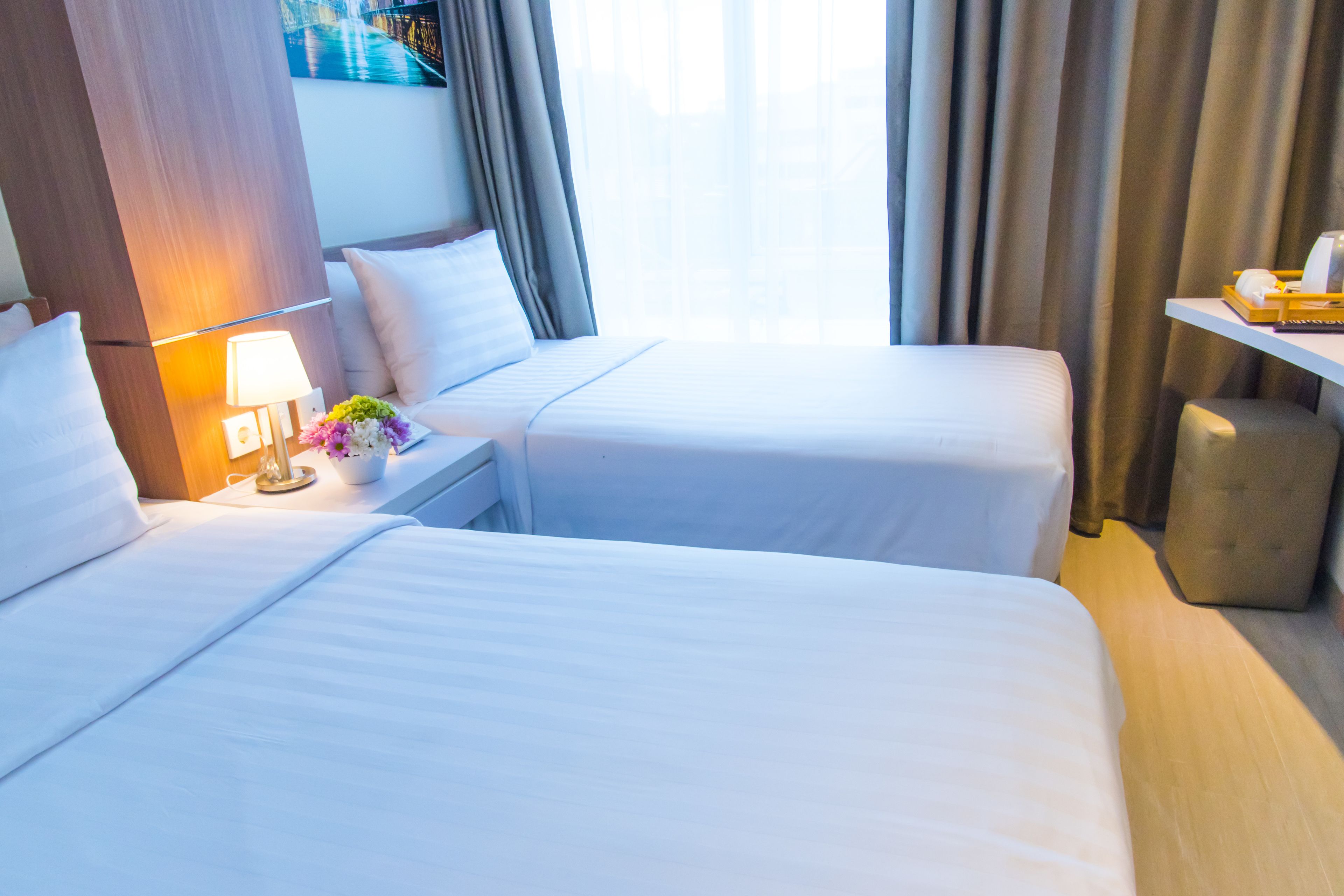 Habitación de hotel con dos camas, una mesita de noche con lámpara, una pequeña mesa con un arreglo floral y vistas a la ciudad a través de amplios ventanales.