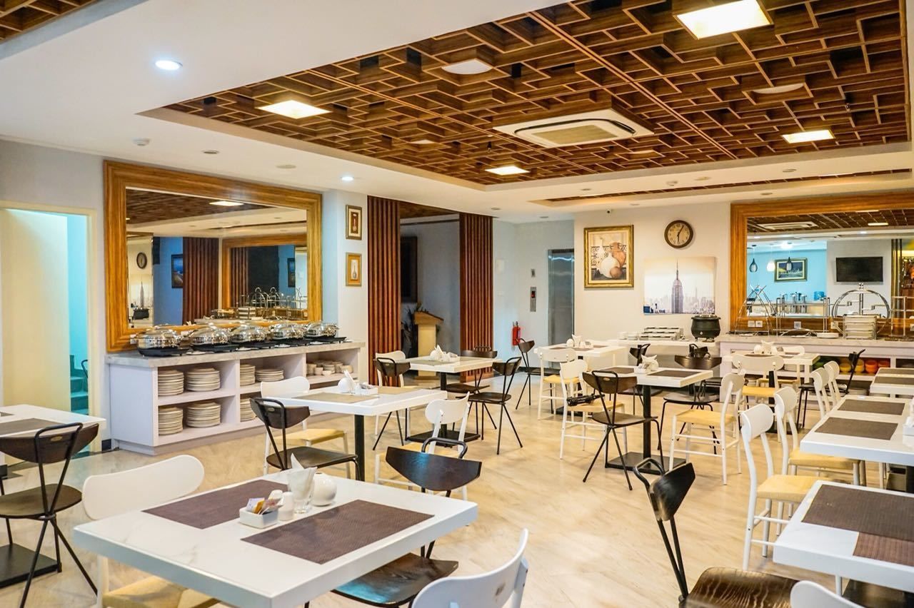 Un espace de restauration moderne, doté de nombreuses tables et chaises, d’un buffet central où sont présentés les plats, et d’un plafond orné d’un motif géométrique.