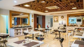 Un espace de restauration moderne, doté de nombreuses tables et chaises, d’un buffet central où sont présentés les plats, et d’un plafond orné d’un motif géométrique.