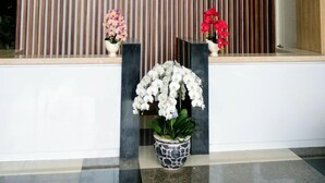 Un área de recepción con un moderno muro de paneles de madera, una orquídea blanca en maceta y dos arreglos florales más pequeños sobre pedestales de mármol.