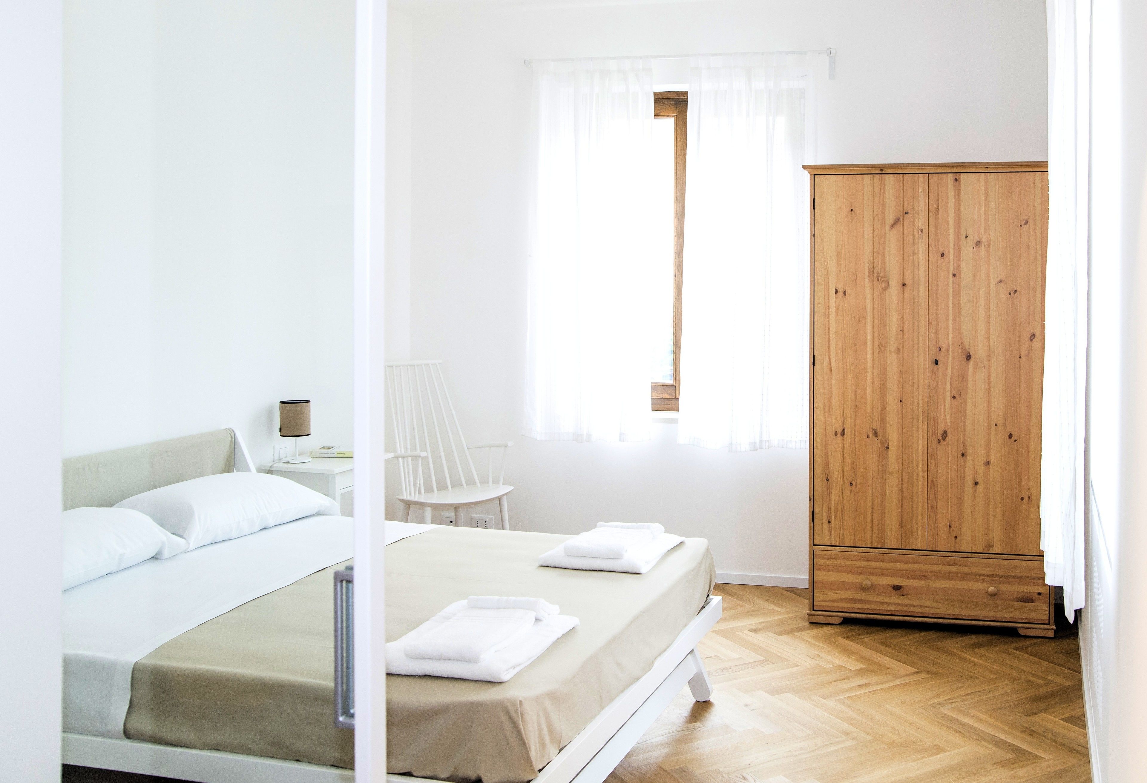Un dormitorio con cama, armario de madera, una silla y una ventana con cortinas.