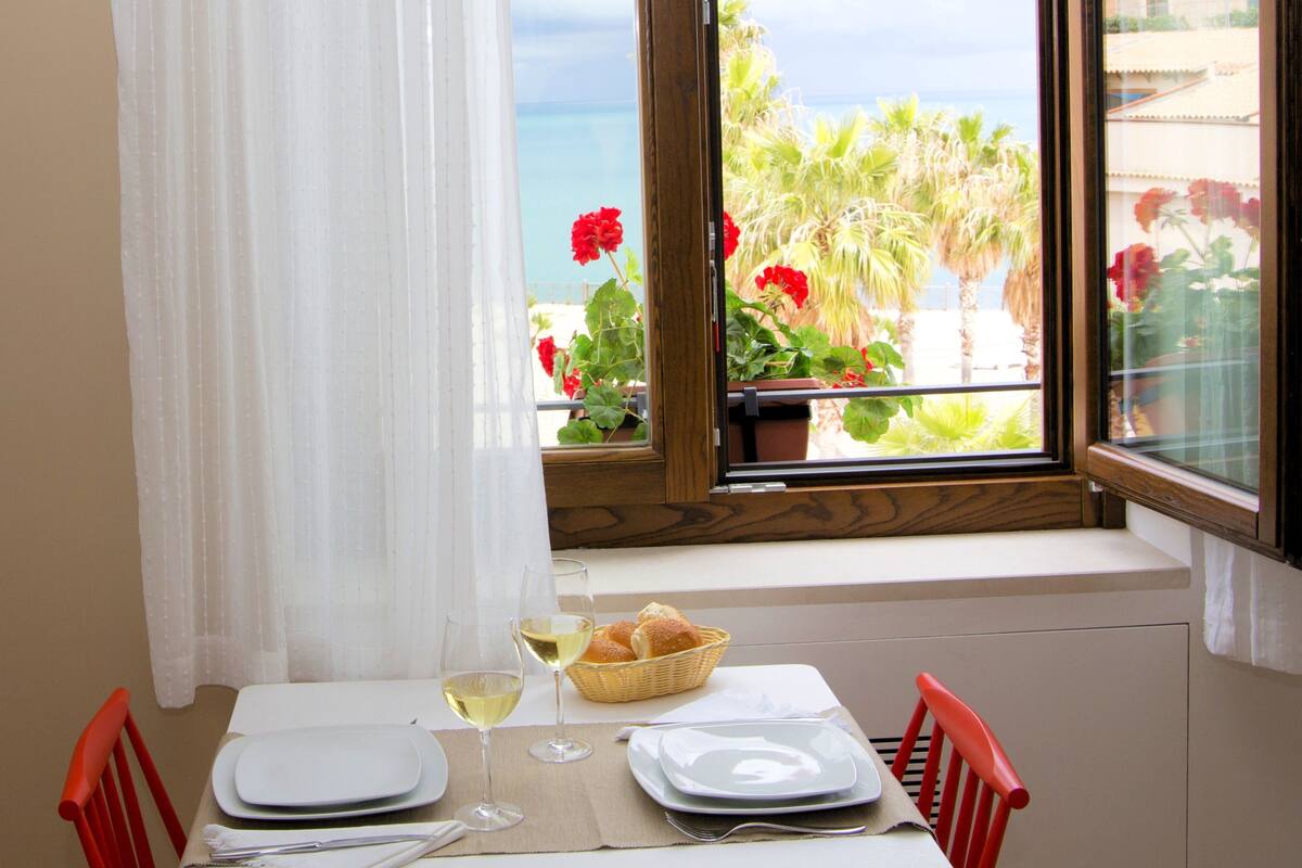 Une table à manger dressée avec des assiettes, des verres et un panier de pain, donnant sur un balcon avec vue sur la mer et les palmiers.