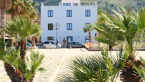 A building with a white facade and multiple windows, surrounded by palm trees and a mountainous backdrop. - Residence ITACA (Castellammare del Golfo)