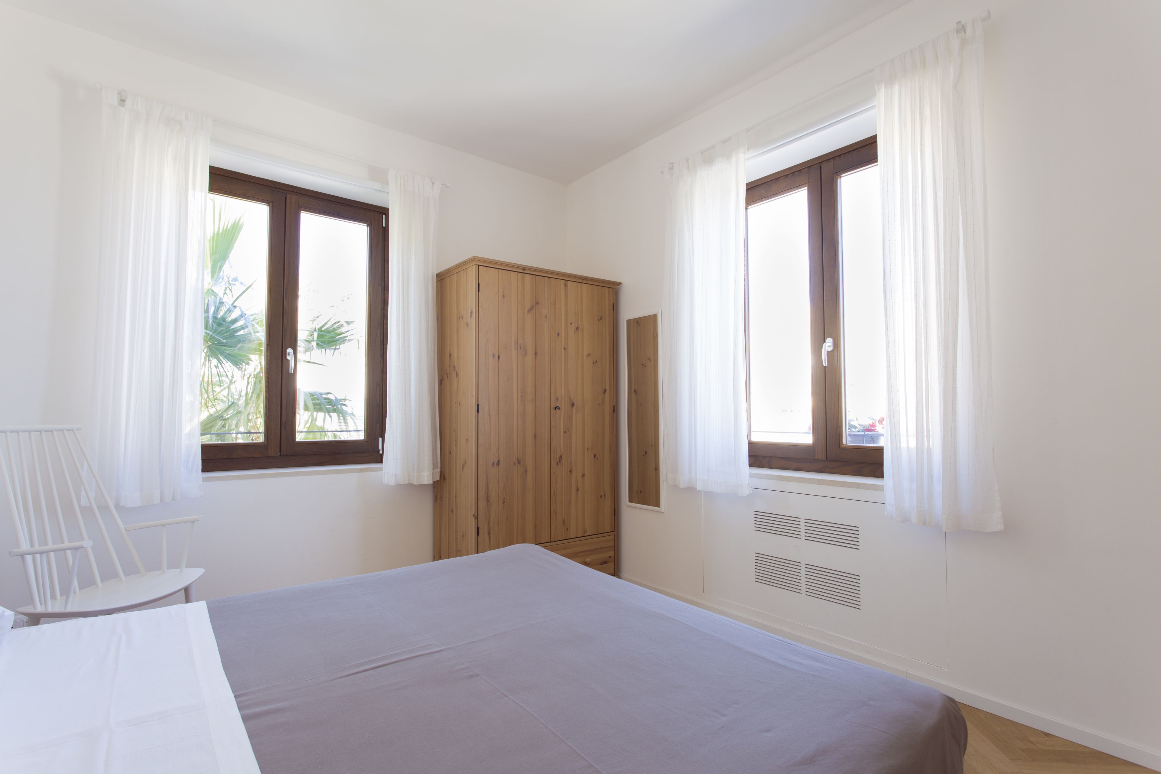 Un dormitorio con cama, un armario de madera y dos ventanas con cortinas blancas.