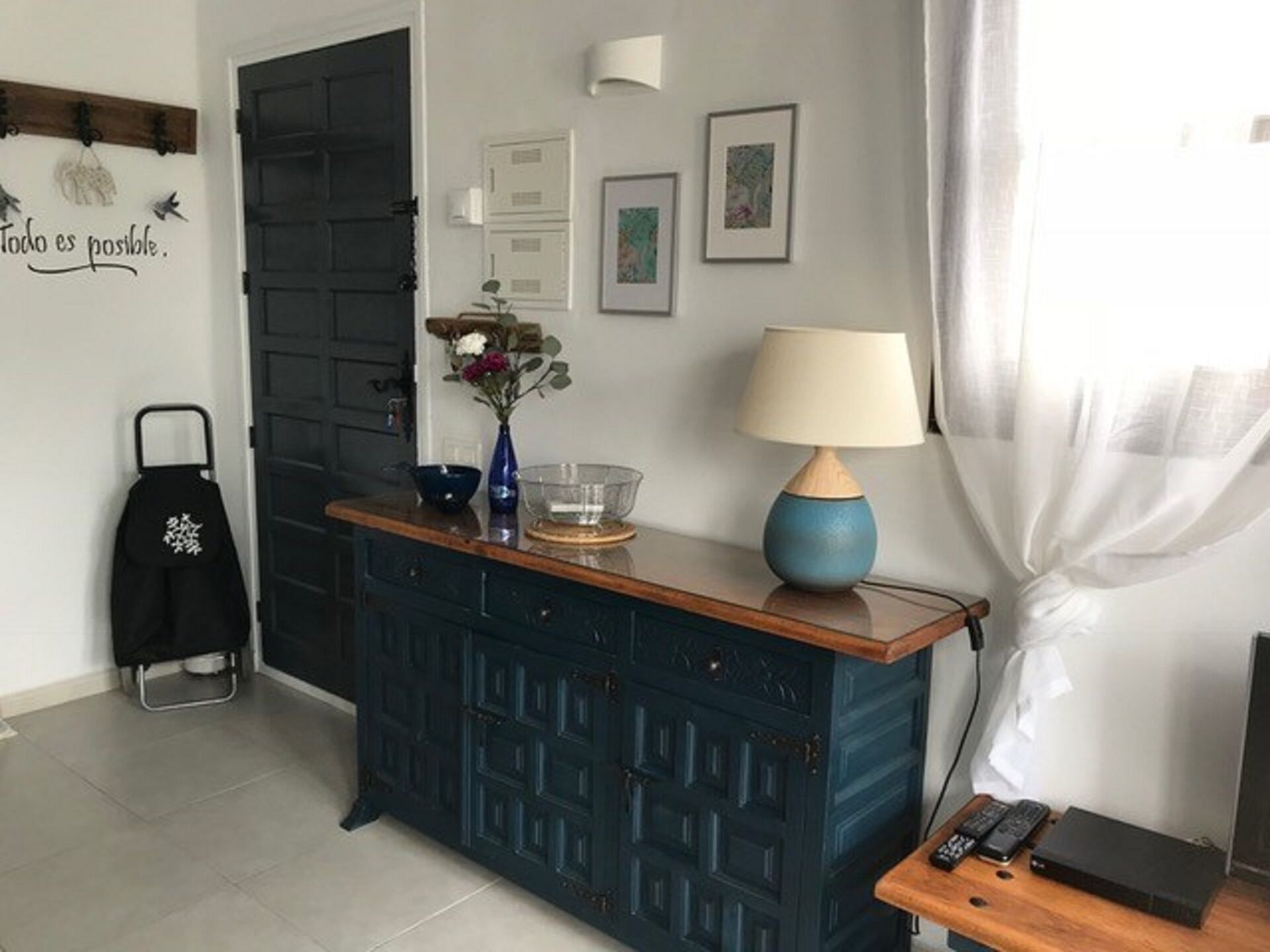 A room with a dark blue cabinet, a small table with a lamp, a bowl, and a vase with flowers. There is a coat rack with a black coat and a wall with a motivational quote.